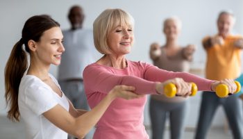 Young,Lady,Coach,Helping,Senior,Woman,While,Exercising,With,Dumbbells,